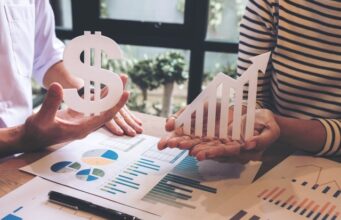 La televisión lineal cae un 2,8% mientras los medios digitales concentran el mayor repunte de inversión publicitaria Dos personas sostienen un símbolo del dólar y una flecha ascendente sobre una mesa con gráficos e informes financieros, representando el crecimiento económico y la evolución de los ingresos.
