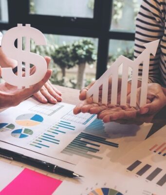 La televisión lineal cae un 2,8% mientras los medios digitales concentran el mayor repunte de inversión publicitaria Dos personas sostienen un símbolo del dólar y una flecha ascendente sobre una mesa con gráficos e informes financieros, representando el crecimiento económico y la evolución de los ingresos.