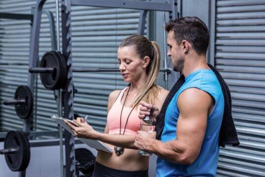 Mujer y hombre conversando junto a una máquina de pesas en un gimnasio mientras revisan una tabla de entrenamiento.