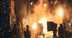 Rodaje cinematográfico con efectos especiales de explosiones y chispas, mientras el equipo técnico trabaja entre cámaras, focos y medidas de seguridad en un set de filmación nocturno.