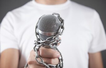 La Agrupación de Periodistas de UGT rechaza la expulsión de los equipos de RTVE y EFE de Castillejos y exige garantías para el libre ejercicio del periodismo Mano que sostiene un micrófono encadenado, símbolo de las restricciones al libre ejercicio del periodismo tras la expulsión de RTVE y EFE de Castillejos.