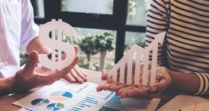 Dos personas sostienen un símbolo del dólar y una flecha ascendente sobre una mesa con gráficos e informes financieros, representando el crecimiento económico y la evolución de los ingresos.