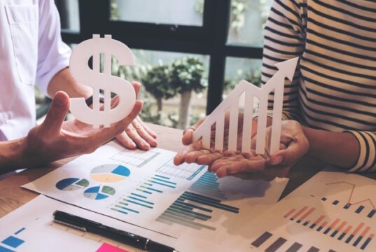 La televisión lineal cae un 2,8% mientras los medios digitales concentran el mayor repunte de inversión publicitaria Dos personas sostienen un símbolo del dólar y una flecha ascendente sobre una mesa con gráficos e informes financieros, representando el crecimiento económico y la evolución de los ingresos.