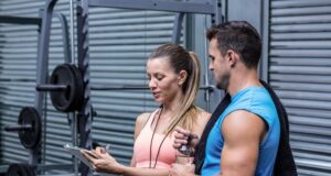 Mujer y hombre conversando junto a una máquina de pesas en un gimnasio mientras revisan una tabla de entrenamiento.