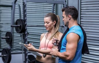 Los usuarios de instalaciones deportivas priorizan la salud y la atención personalizada Mujer y hombre conversando junto a una máquina de pesas en un gimnasio mientras revisan una tabla de entrenamiento.