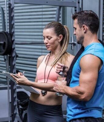 Los usuarios de instalaciones deportivas priorizan la salud y la atención personalizada Mujer y hombre conversando junto a una máquina de pesas en un gimnasio mientras revisan una tabla de entrenamiento.
