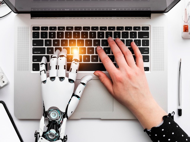 Impacto laboral de la IA representado por una mano humana y una mano robótica trabajando juntas sobre el teclado de un ordenador portátil.