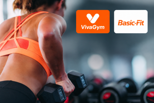 Mujer entrenando con pesas en un gimnasio junto a los logos de VivaGym y Basic-Fit, dos de las cadenas que protagonizan las fusiones en el sector de instalaciones deportivas y gimnasios en España.