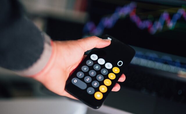 Mano con un móvil que muestra una calculadora, frente a un ordenador; imagen alusiva al cálculo del IVA.
