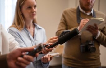 La UE abre 20,5 millones de euros en ayudas al periodismo y al pluralismo Periodista sostiene un micrófono durante una entrevista mientras otro reportero toma notas junto a una cámara.