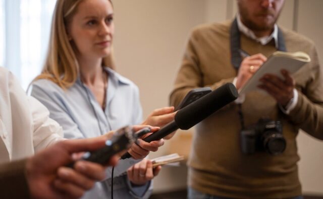 press-reporter-fallowing-leads-case-680x420 Periodista sostiene un micrófono durante una entrevista mientras otro reportero toma notas junto a una cámara.