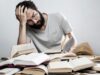 Trabajador con gesto de agotamiento entre libros, imagen que ilustra el estrés y los riesgos psicosociales en el trabajo.