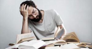 Trabajador con gesto de agotamiento entre libros, imagen que ilustra el estrés y los riesgos psicosociales en el trabajo.