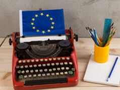 UGT ante la Misión Europea: empleo estable y periodismo de calidad Máquina de escribir roja con una bandera de la Unión Europea, acompañada de un cuaderno y un recipiente con lápices y pinceles sobre una mesa.