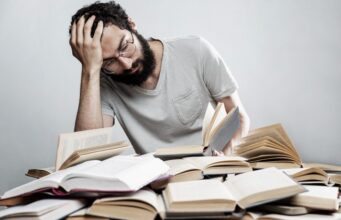 La seguridad laboral también importa en la cultura Trabajador con gesto de agotamiento entre libros, imagen que ilustra el estrés y los riesgos psicosociales en el trabajo.