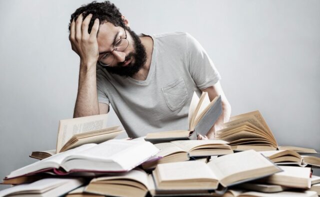 Trabajador con gesto de agotamiento entre libros, imagen que ilustra el estrés y los riesgos psicosociales en el trabajo.