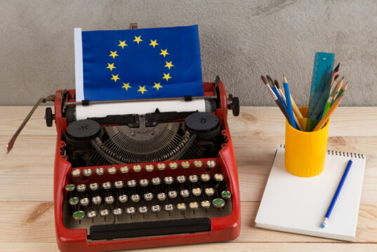 Máquina de escribir roja con una bandera de la Unión Europea, acompañada de un cuaderno y un recipiente con lápices y pinceles sobre una mesa.