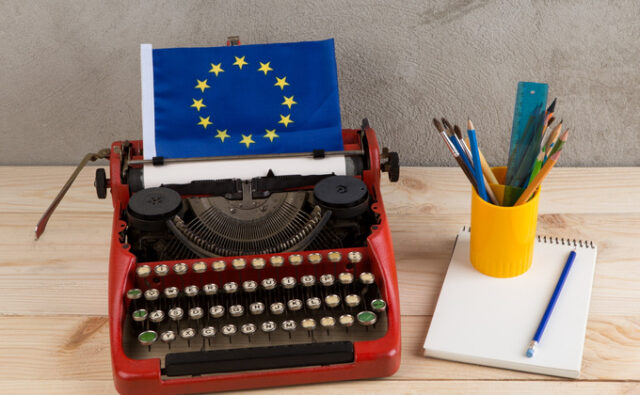 Máquina de escribir roja con una bandera de la Unión Europea, acompañada de un cuaderno y un recipiente con lápices y pinceles sobre una mesa.
